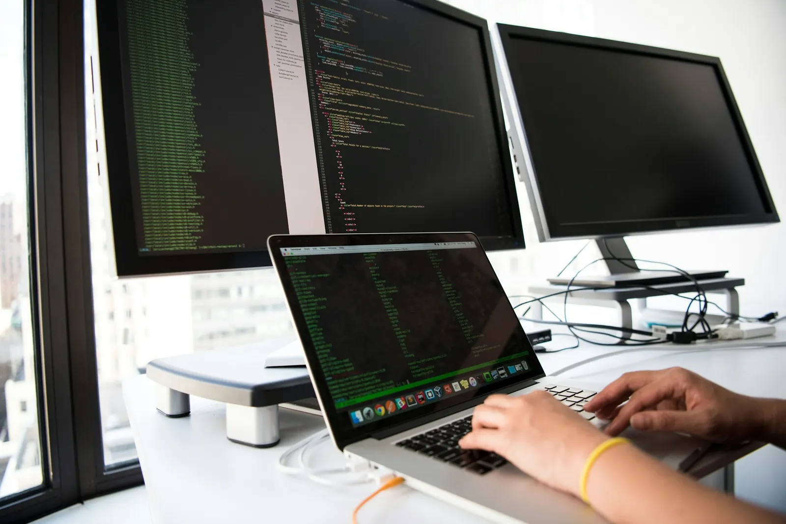 Hands typing on a laptop in front of monitors full of code