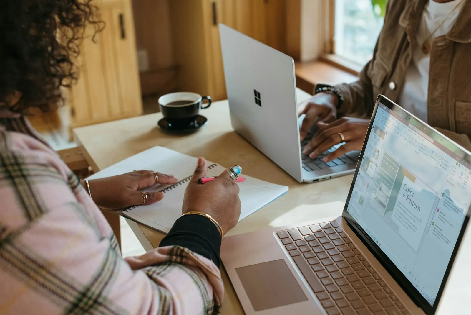 A person taking notes in a notebook beside two open laptops