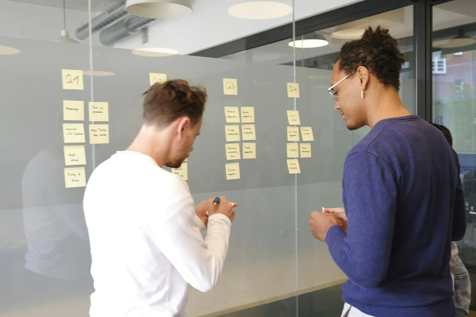 Two people adding sticky notes to a planning board on a glass wall